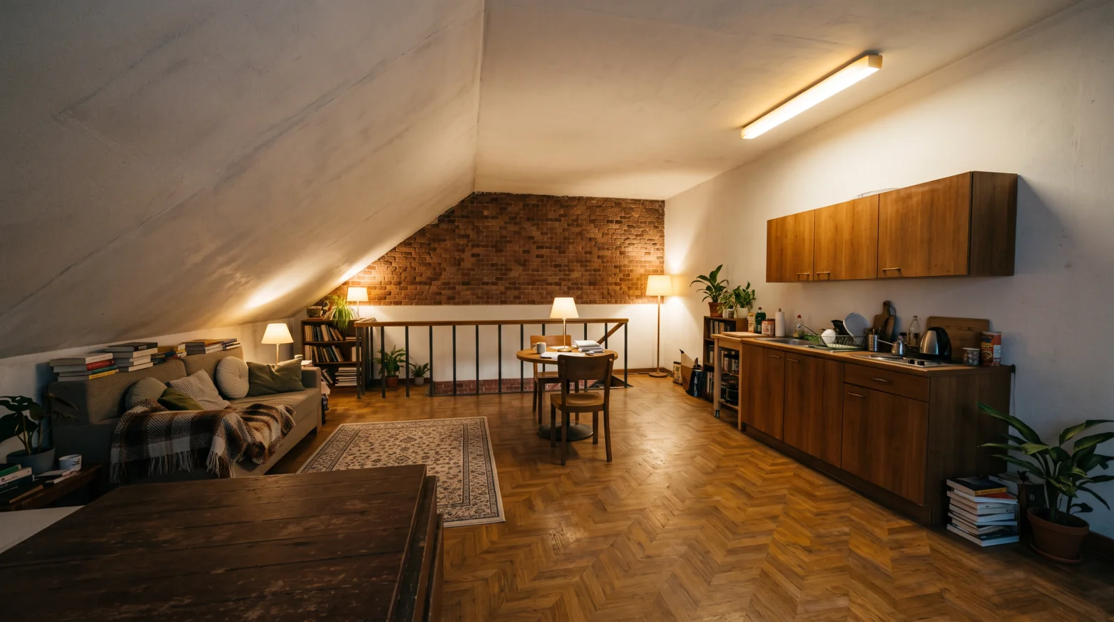 Image d'ambiance générée par IA. Séjour mansardé éclairé par des lampes, plafonds en pente, mur pignon en brique, canapé, table ronde, garde-corps de trémie et kitchenette en bois.